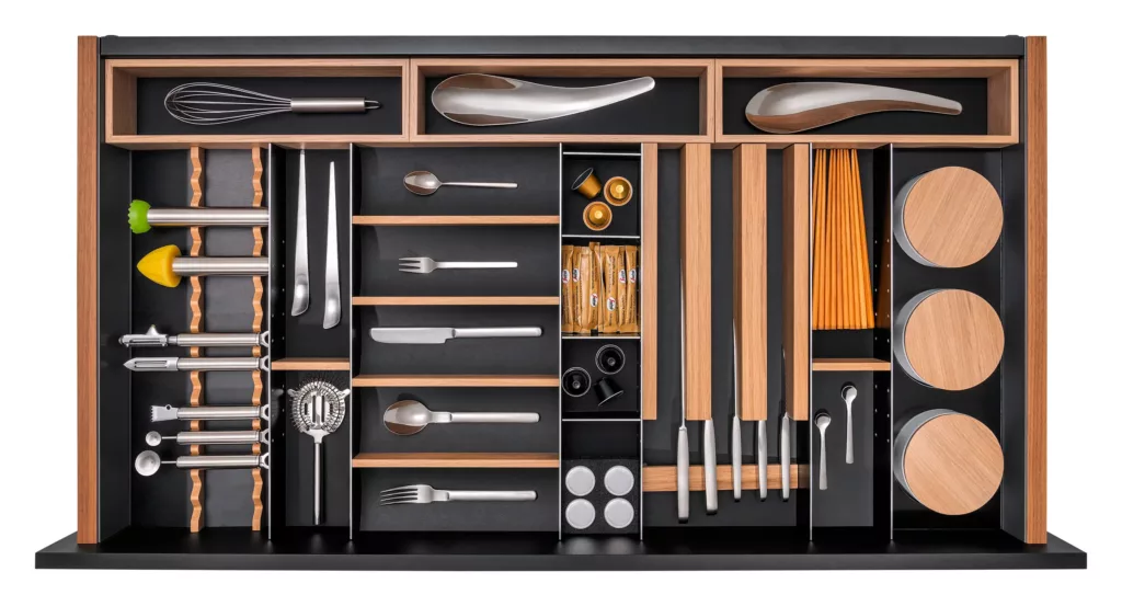 Geordnetes Fach mit Besteck, Schneebesen, Küchenutensilien, Holzstäben und runden Behältern.