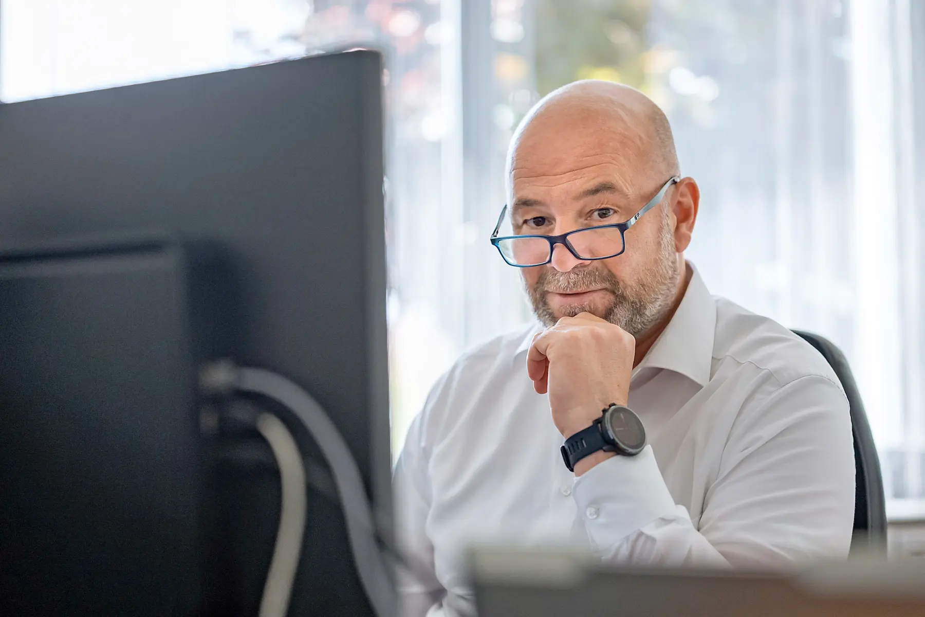 Mann mit Brille und Uhr in weißem Hemd sitzt am Schreibtisch vor einem Computerbildschirm.