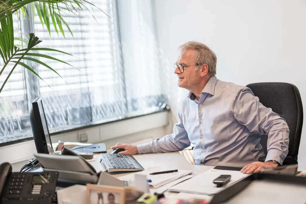 Älterer Mann arbeitet an einem Schreibtisch mit Computer, Büromaterialien und Pflanze im Büro.