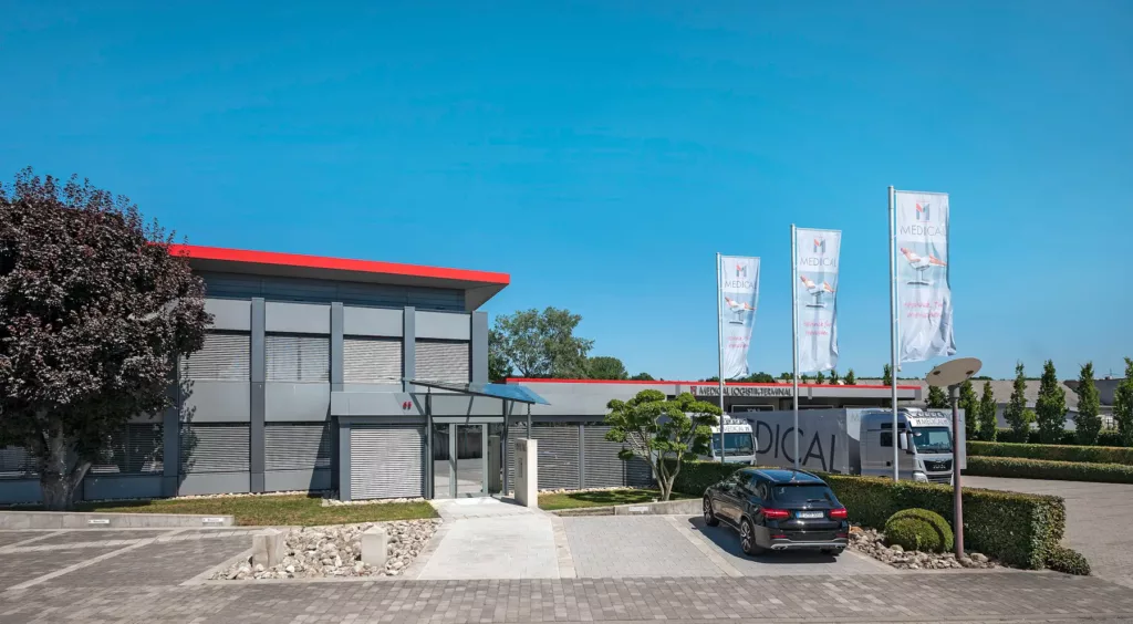 Modernes Gebäude mit grauer Fassade und rotem Dach, umgeben von Parkplatz und Fahnen "Medical".