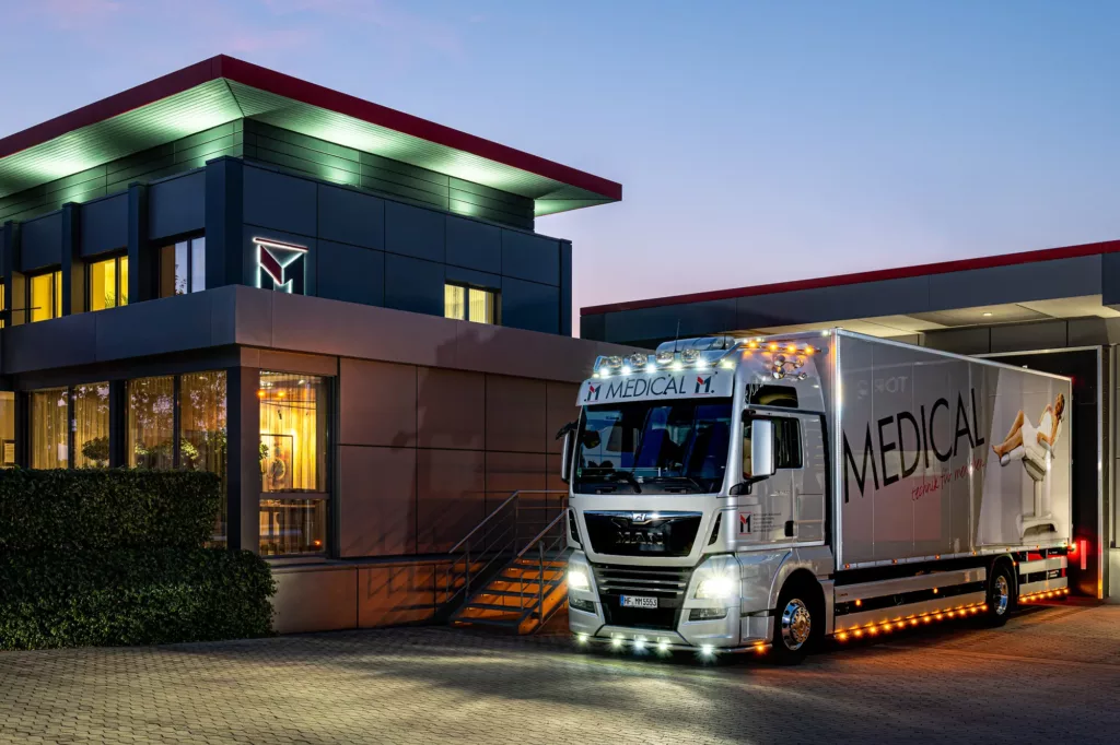 Beleuchteter Lkw mit Aufschrift „MEDICAL“ vor modernem Gebäude in der Abenddämmerung.