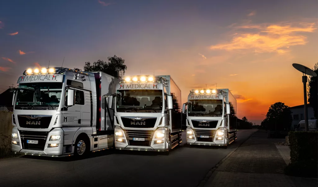 Drei beleuchtete Lkw mit "Medical"-Aufschrift stehen auf einer Straße bei Sonnenuntergang.