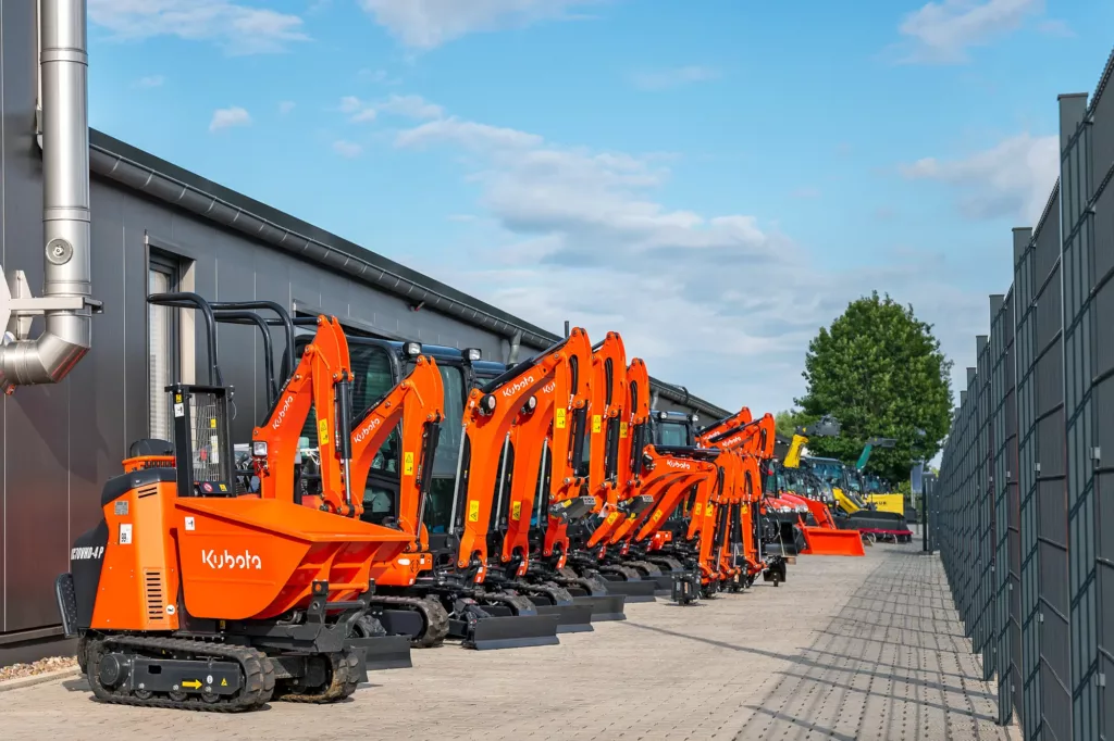Orangefarbene Bagger auf gepflastertem Hof neben Gebäude und Zaun, Himmel leicht bewölkt.