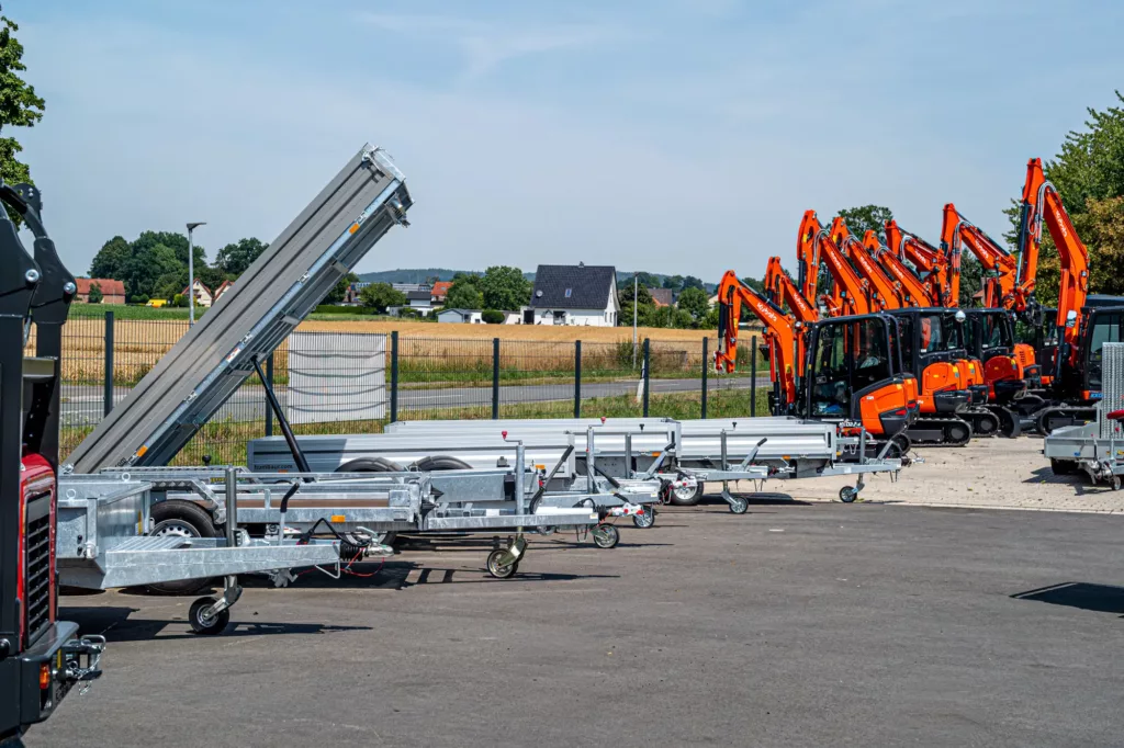 Anhänger und orangene Bagger auf Freigelände, im Hintergrund Landwirtschaft und Gebäude.