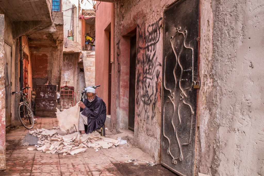 Älterer Mann bearbeitet Lederstücke in enger Gasse mit Graffiti, Fahrrad und Metalltor.