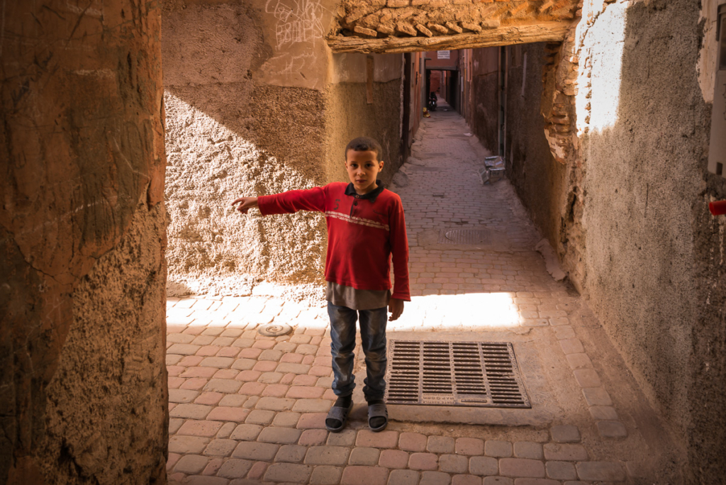 Kind zeigt in schmalem, gepflasterten Durchgang zwischen erdfarbenen Wänden in eine Richtung.