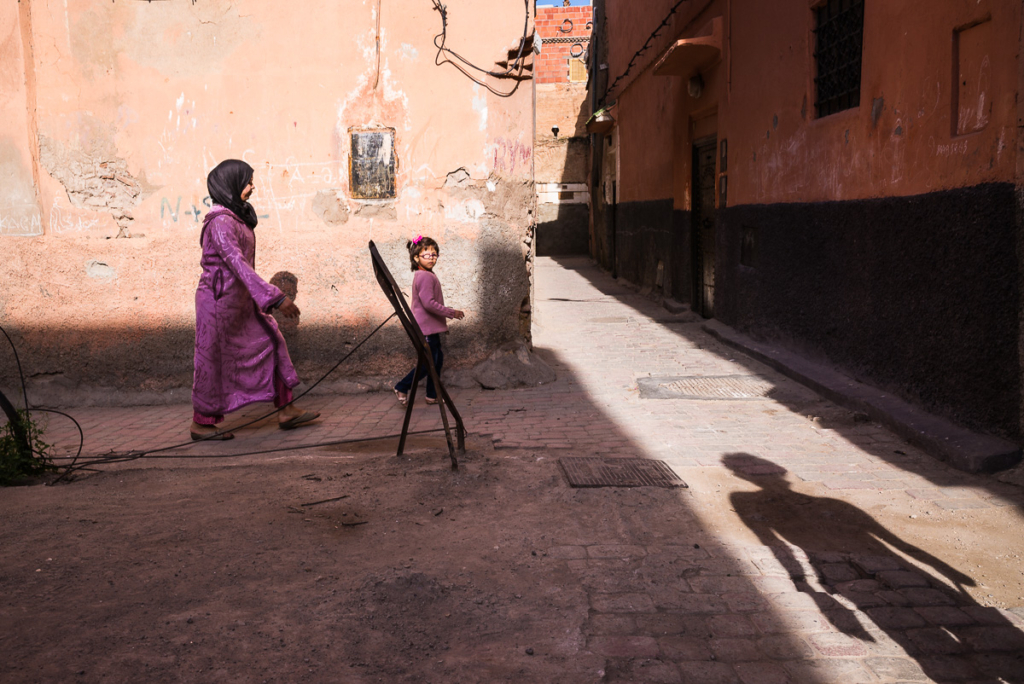 Frau und Mädchen in einer von Licht und Schatten geprägten, erdfarbenen Gasse mit Metallgestell.