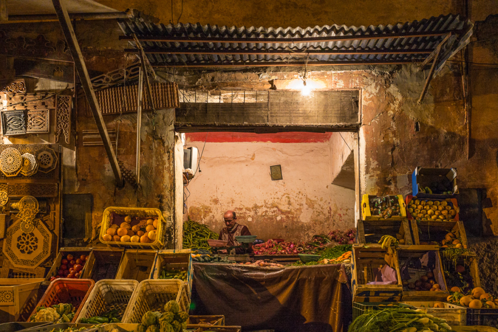 Eingeschlossener Gemüsestand mit Verkäufer, beleuchtet von Glühbirne, mit Schnitzereien.