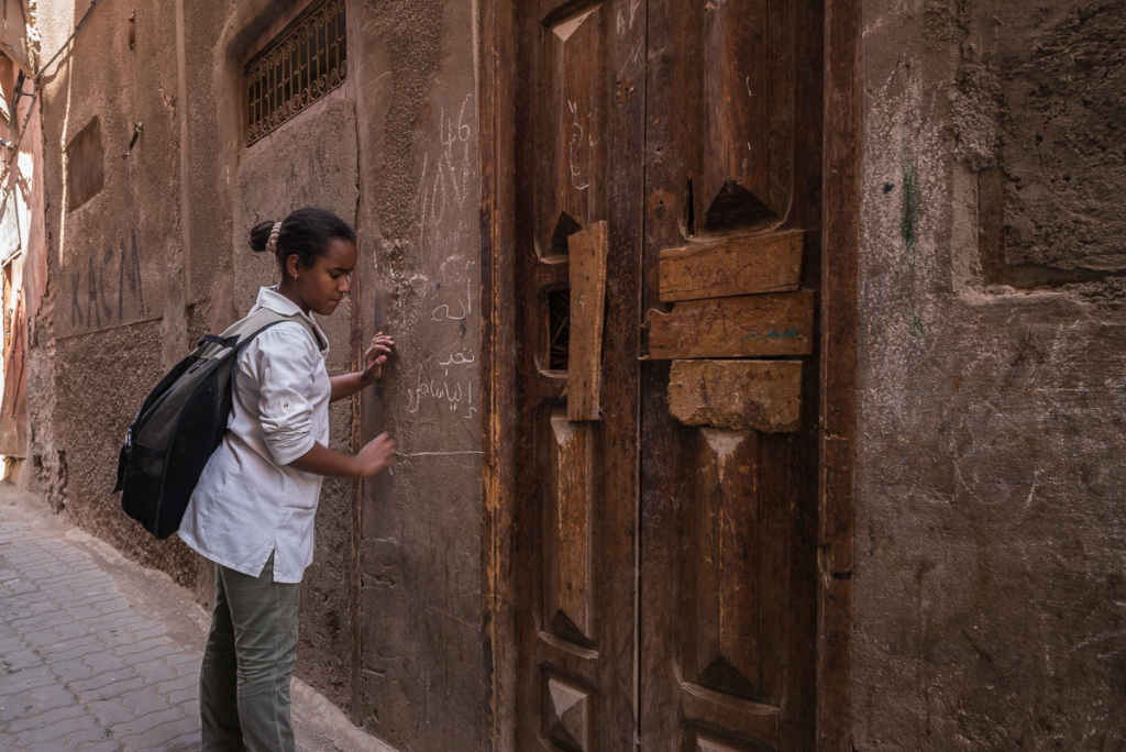 Person schreibt mit Kreide an die Wand, steht in enger Gasse vor abgenutzter Holztür.