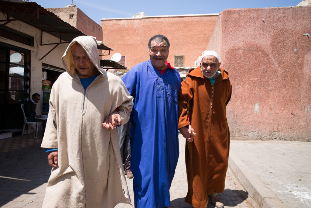 Drei Männer in traditionellen Kleidungen auf einer städtischen Straße, zwei halten Händchen.