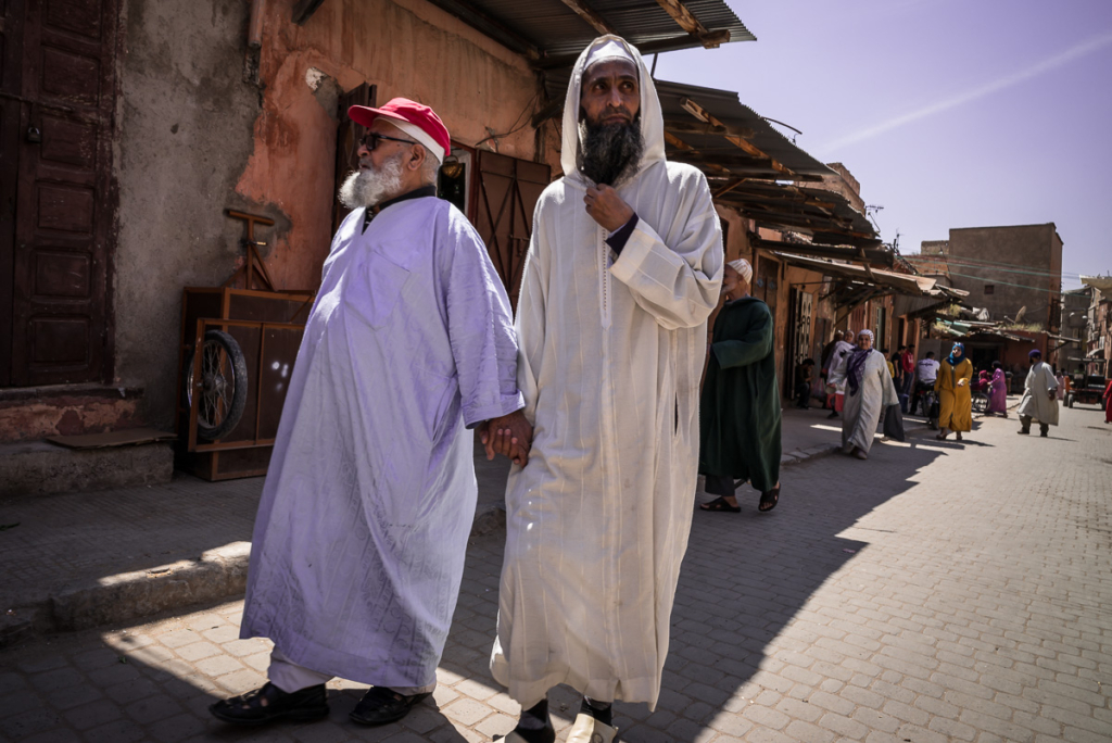 Zwei Männer in traditioneller Kleidung gehen Hand in Hand auf einer sonnigen, belebten Straße.
