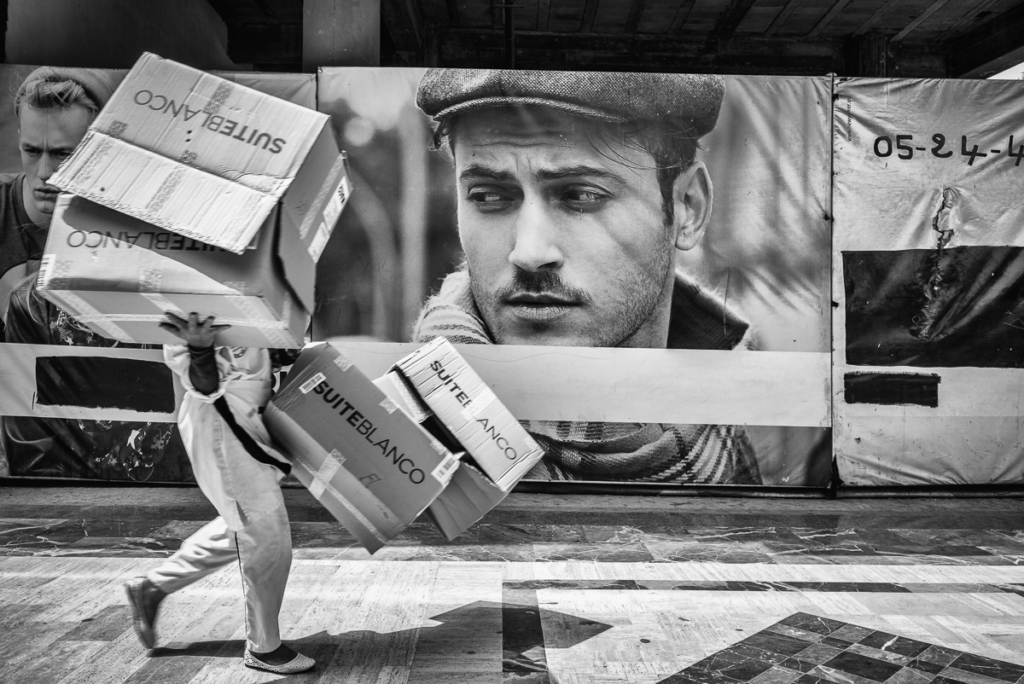 Person trägt Kartons; im Hintergrund große Männerporträt-Werbung auf Plakatwand.