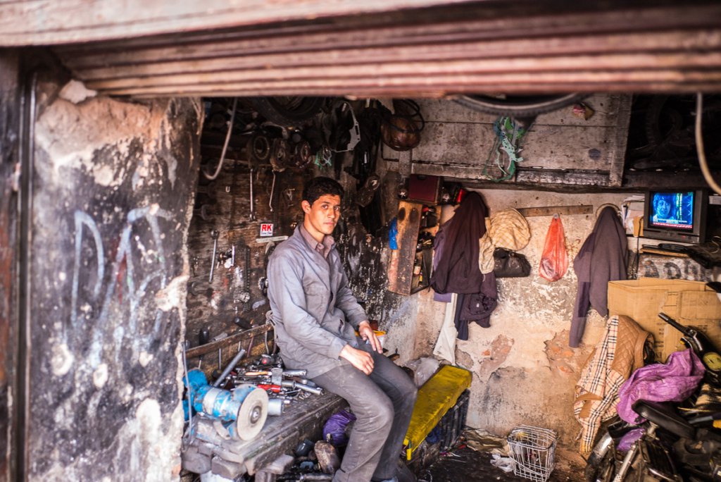 Mann in Werkstatt auf Werkbank mit Werkzeugen, abgenutzte Wände, Kleidung, Tüte, TV im Hintergrund.