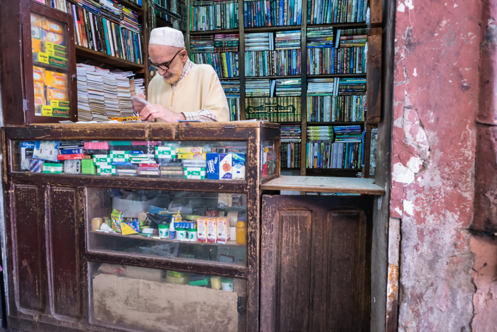 Älterer Mann in traditioneller Kleidung hinter Buchladentresen mit vollen Regalen und Vitrine.