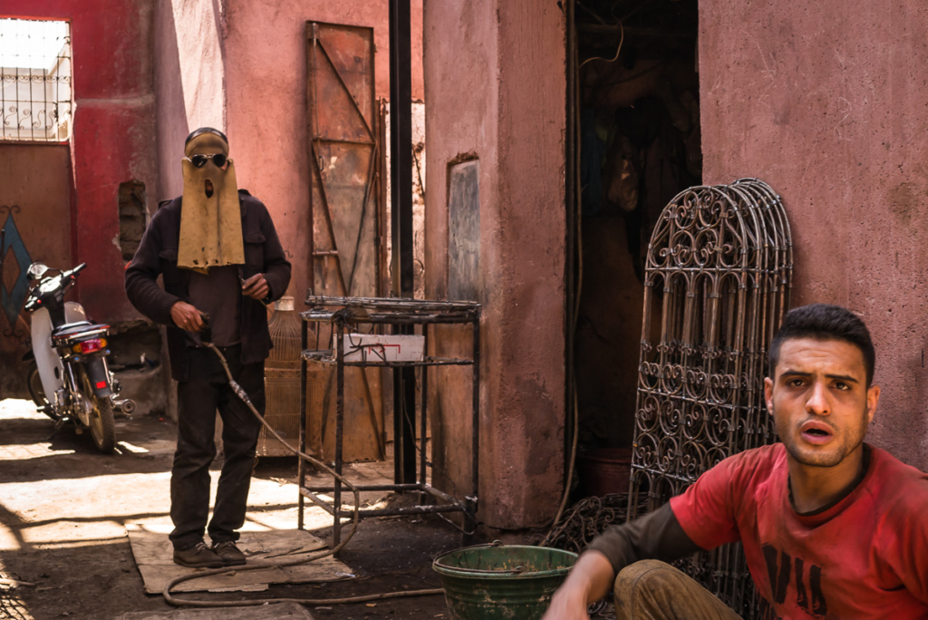 Mann mit Schutzbrille neben Motorrad in Gasse, anderer Mann im Vordergrund vor Metallgitter.