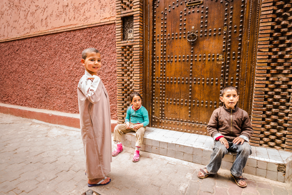 Drei Kinder vor einer verzierten Holztür in einem Innenhof mit gemusterten Wänden.