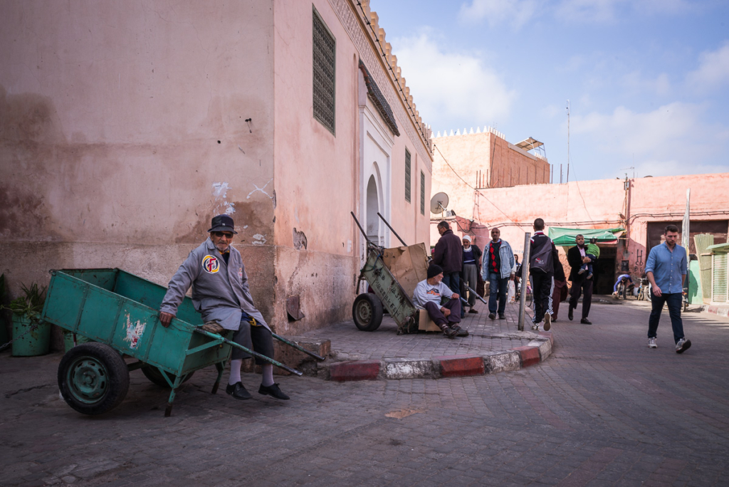 Straßenszene mit Menschen, Handkarren und rosa Gebäude unter bewölktem Himmel.