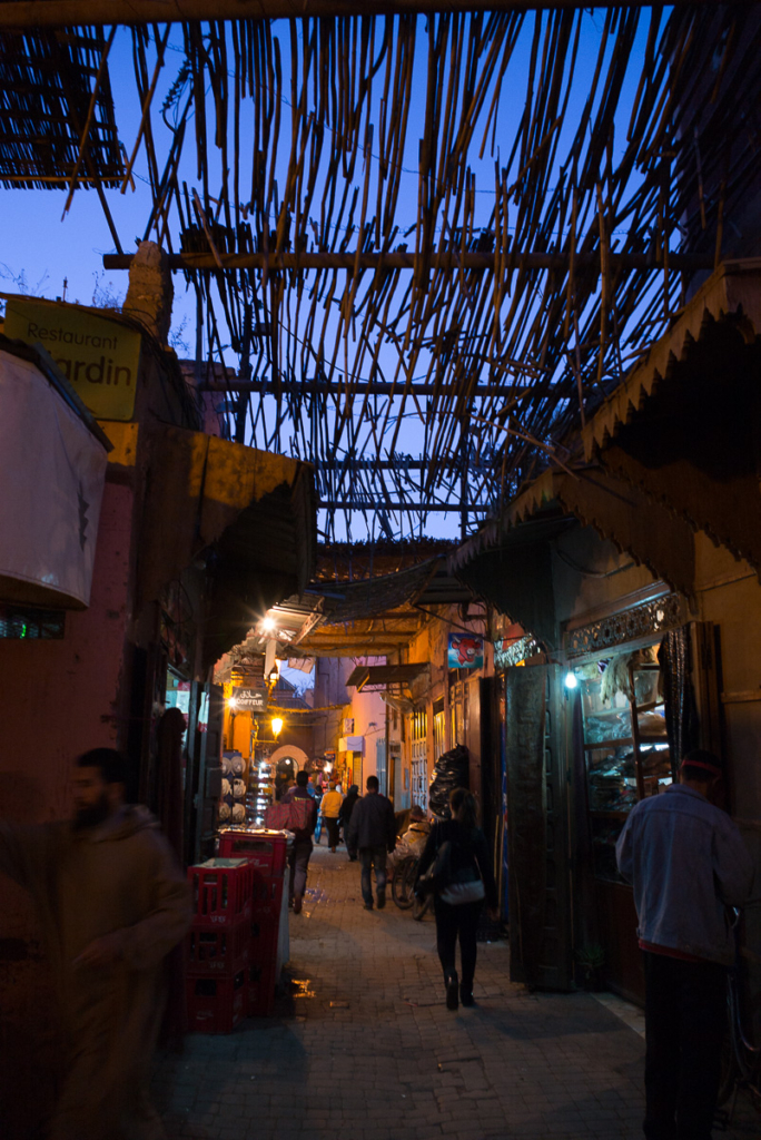 Belebte Marktstraße am Abend, beleuchtete Geschäfte, unregelmäßiges Stangendach, blauer Himmel.