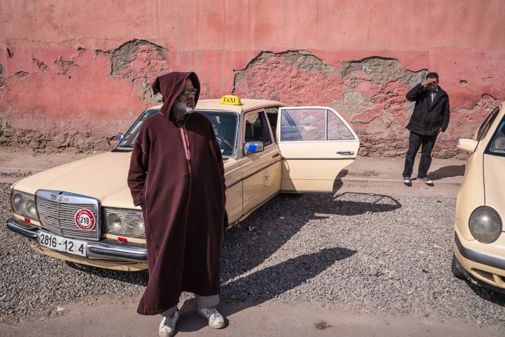 Älterer Mann vor beigem Taxi, andere Person schaut entfernt, beschädigte Wand im Hintergrund.
