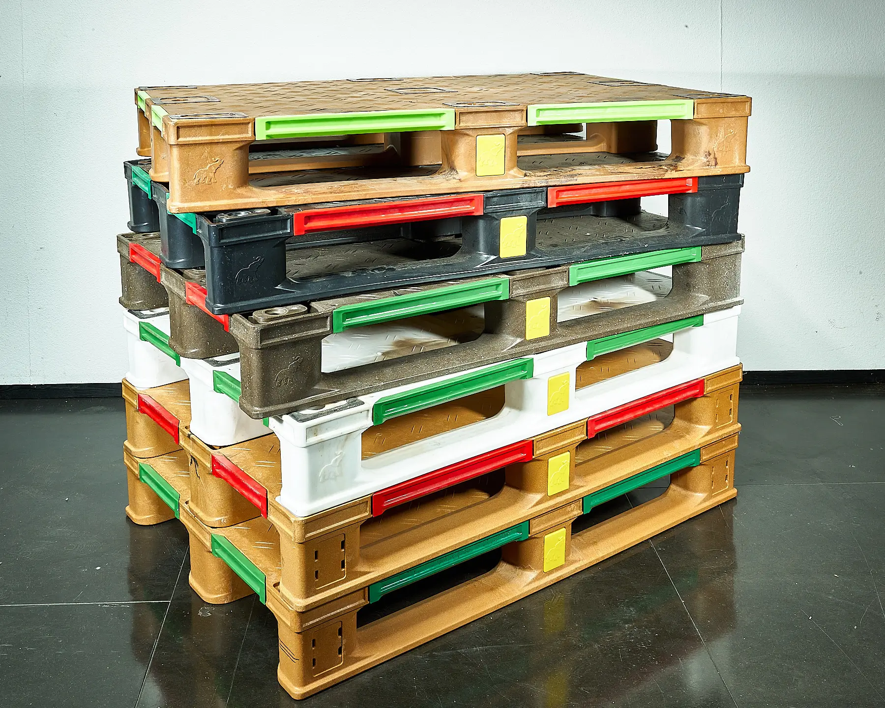 Stapel bunter Kunststoffpaletten auf schwarzem Boden vor weißer Wand.