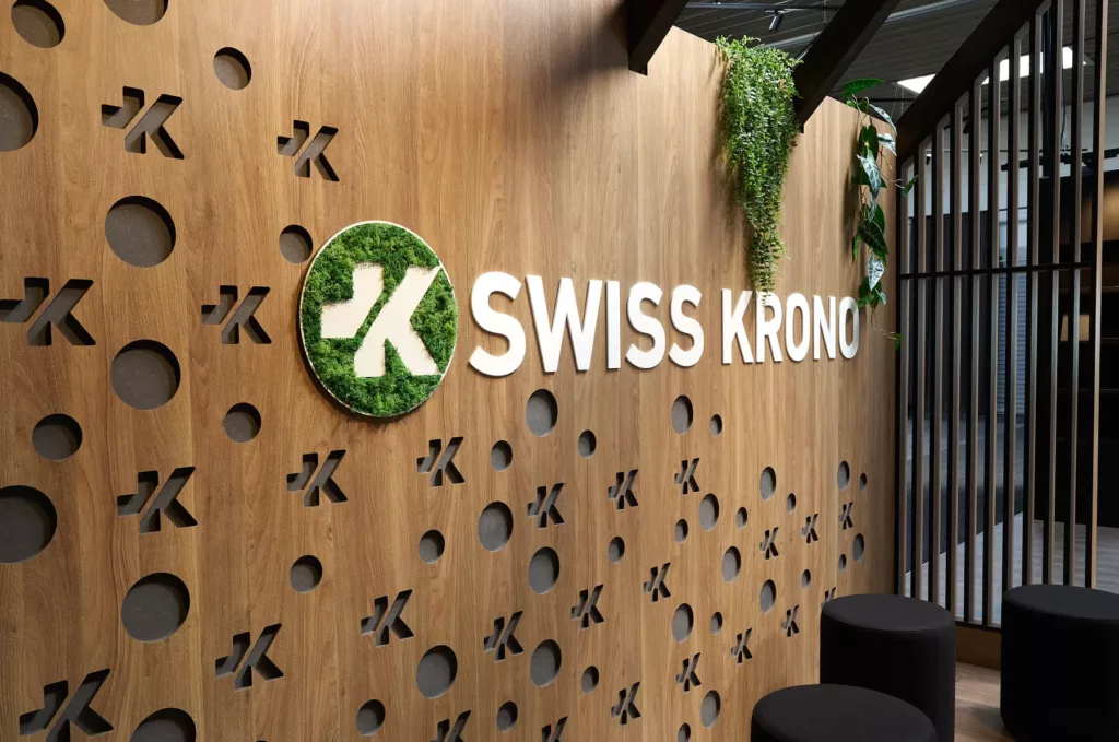 Holzwand mit SWISS KRONO-Logo, runde Ausschnitte und Pflanzen als Dekor.