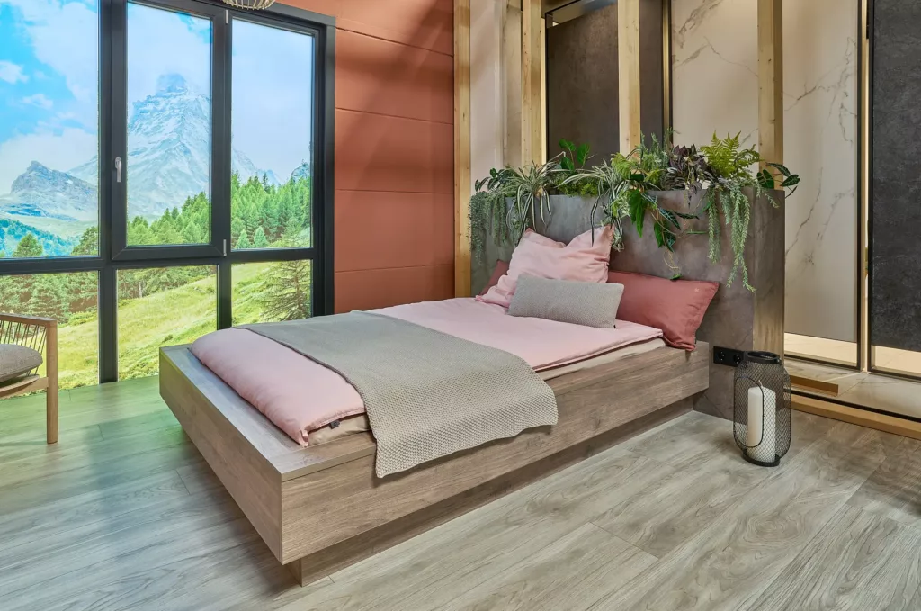 Modernes Schlafzimmer mit Holzfußboden, Bett und Pflanzen. Fensterblick auf grüne Landschaft.