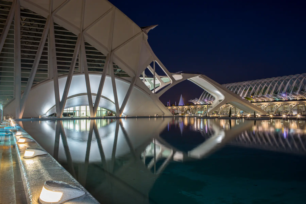 Modernes, beleuchtetes Bauwerk bei Nacht, reflektiert auf Wasser, mit geschwungenen Elementen.