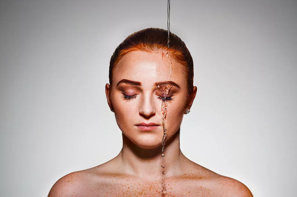Frau mit geschlossenen Augen, Wasserstrahl fließt über ihr Gesicht, einfarbiger Hintergrund.