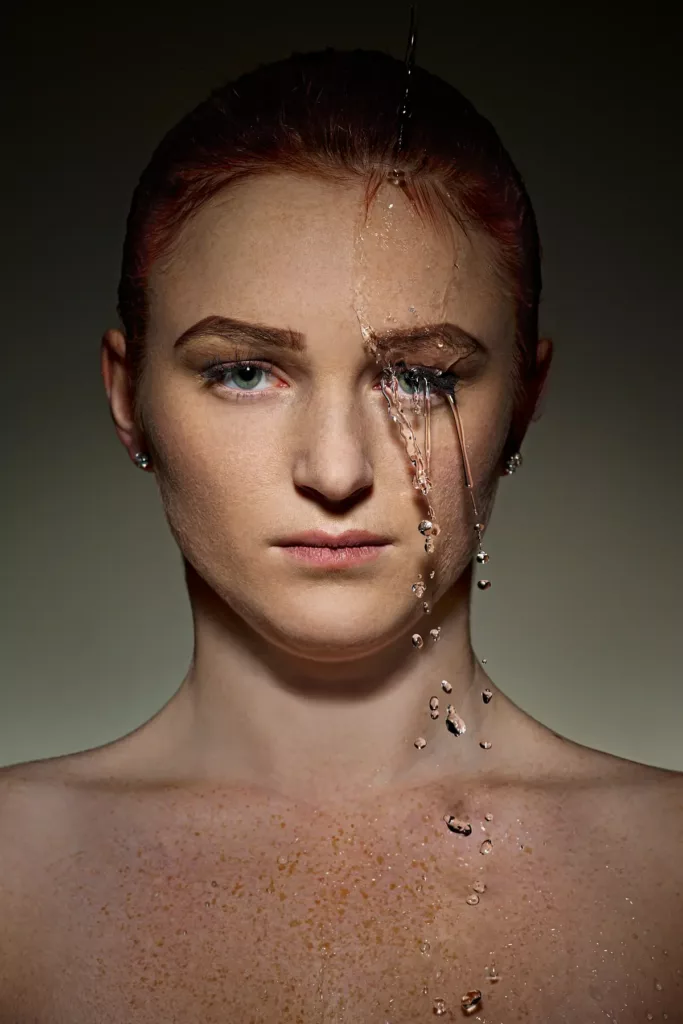 Frau mit frecklem Gesicht, rotem Haar, Wasser tropft über Auge, dunkler Hintergrund.