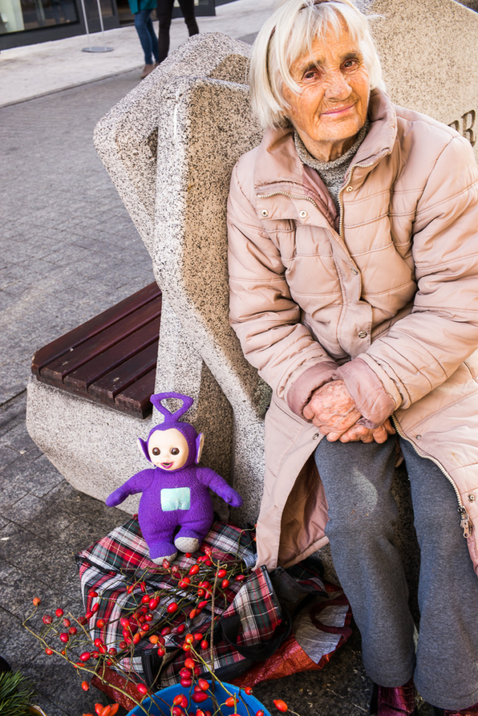Ältere Frau auf Bank mit lila Stofftier und Korb voller roter Früchte.