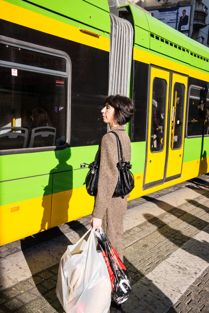 Frau mit Einkaufstüten an Haltestelle neben gelb-grüner Straßenbahn.