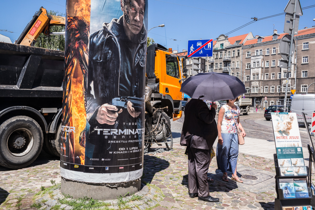 Mann mit Regenschirm und Frau an Litfaßsäule mit Filmplakat, orangefarbener LKW im Hintergrund.