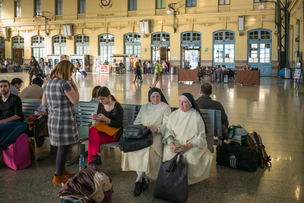 Bahnhofswartesaal mit Menschen auf Bänken, darunter zwei Nonnen in traditionellen Gewändern.
