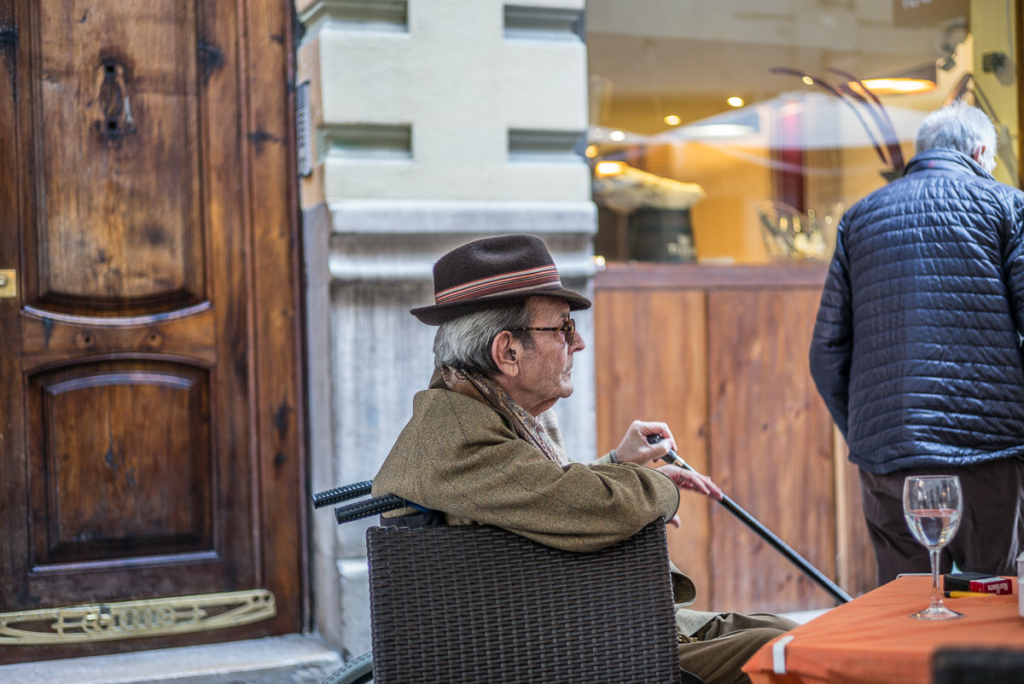 Älterer Mann mit Hut und Gehstock sitzt draußen, leerer Stuhl, Weinglas, Passant im Hintergrund.
