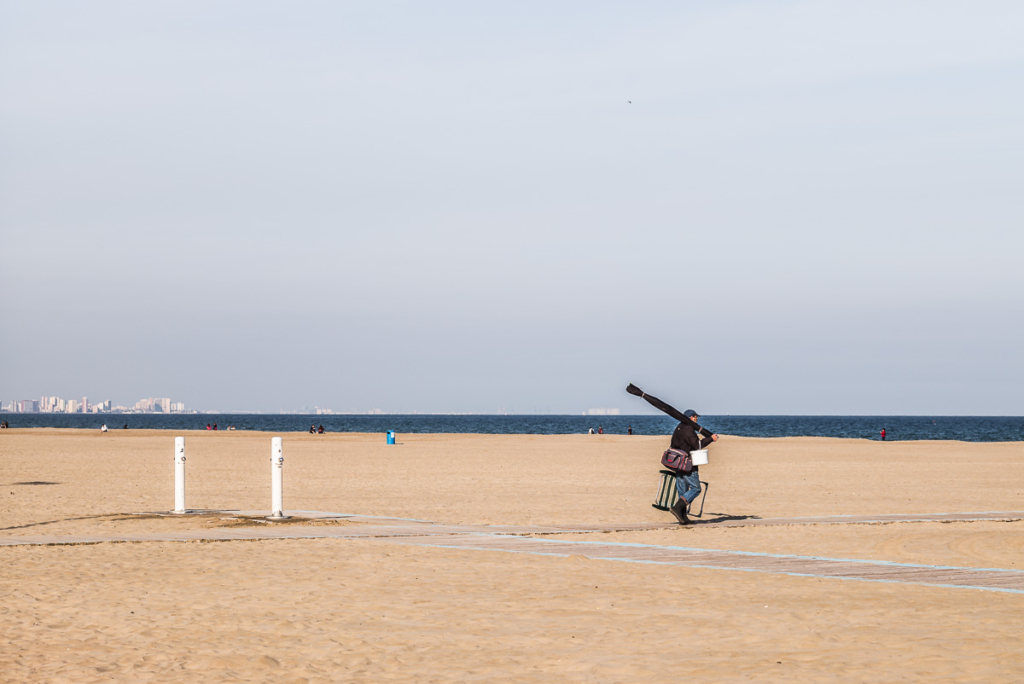 Person am leeren Sandstrand mit Angelausrüstung, Meer und ferne Stadt im Hintergrund.