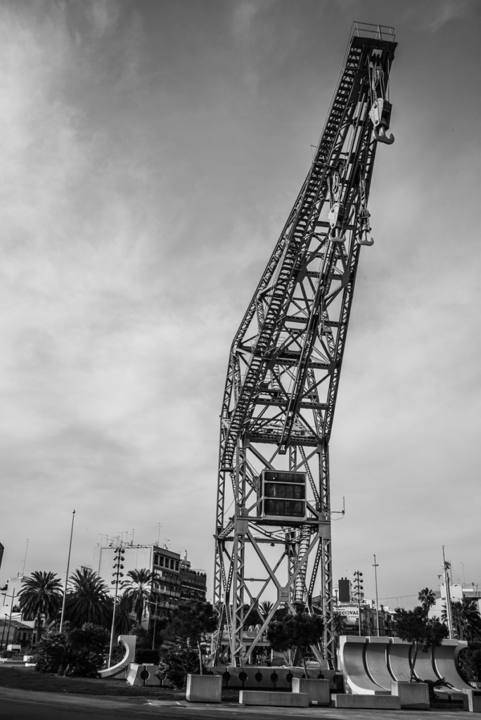 Großer Metallkran in urbaner Umgebung mit Palmen und modernen Gebäuden unter bewölktem Himmel.