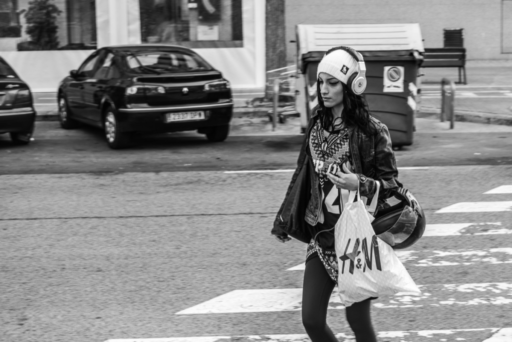 Person mit Kopfhörern, Einkaufstasche und Helm überquert Zebrastreifen. Autos im Hintergrund.