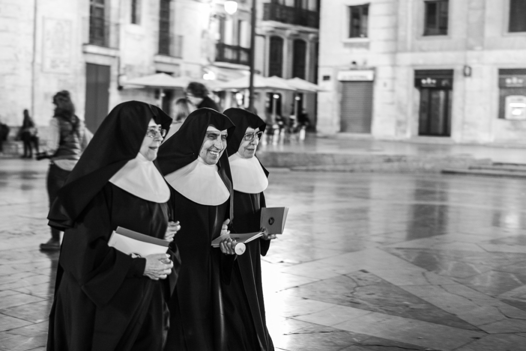 Drei Nonnen in traditionellen Gewändern mit Büchern in beleuchteter städtischer Umgebung.