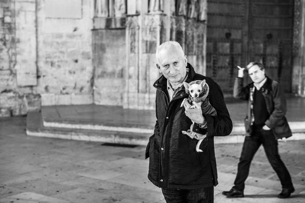 Älterer Mann mit Hund, anderer Mann im Hintergrund gestikuliert, architektonisch interessanter Ort.