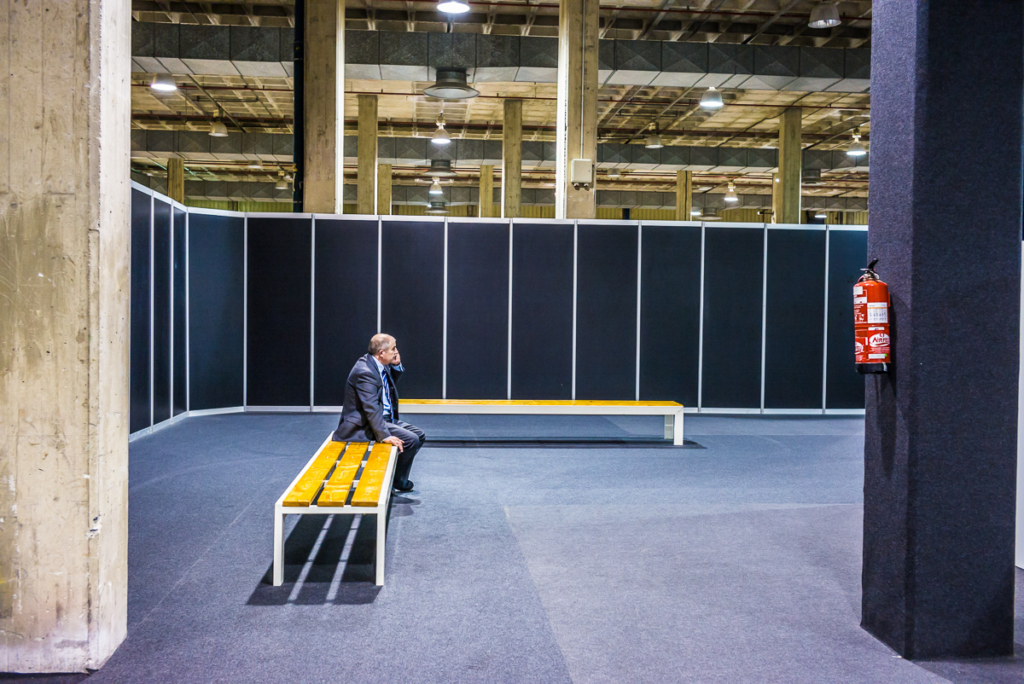 Person sitzt auf gelber Bank in großer Halle mit schwarzen Wänden und Feuerlöscher an der Wand.
