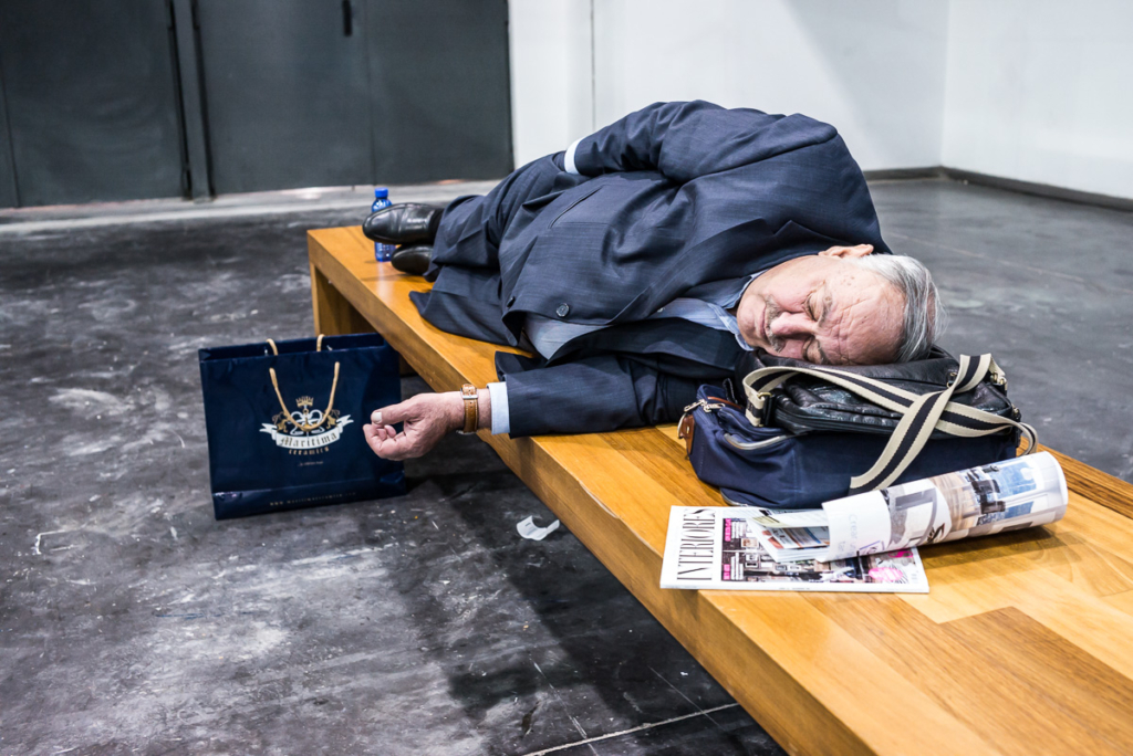 Mann im Anzug schläft auf Bank mit Tasche, Zeitschrift und Einkaufstüte daneben.
