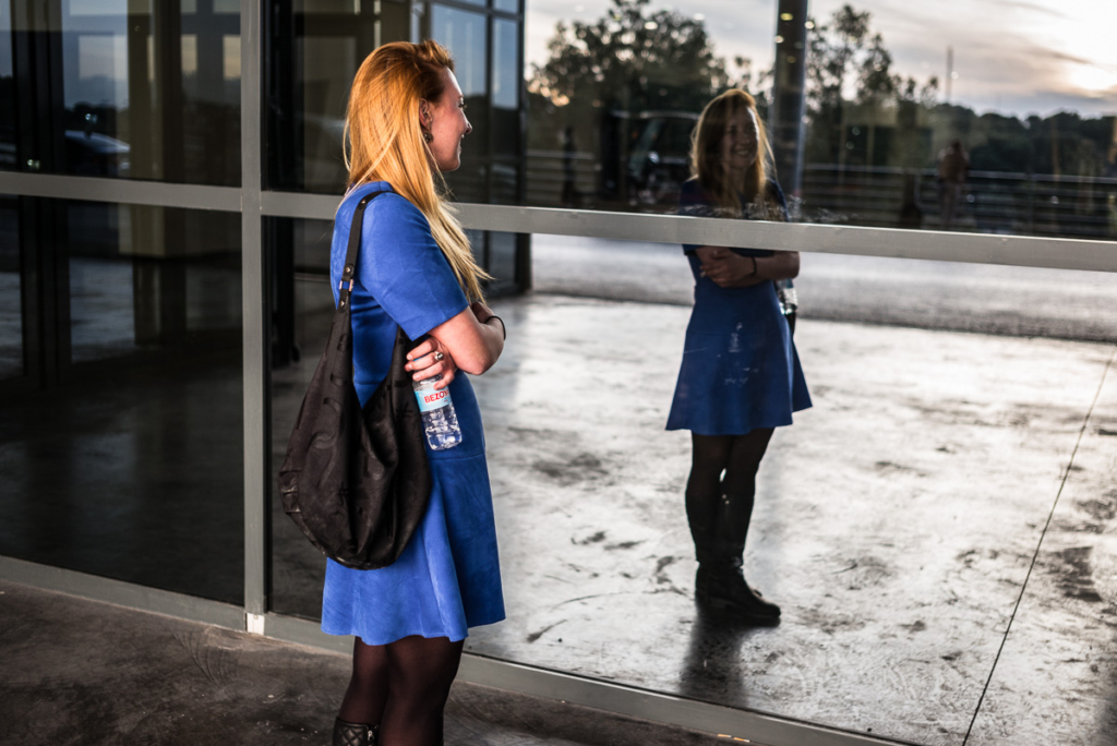 Frau im blauen Kleid vor Glaswand mit schwarzer Tasche und Wasserflasche.