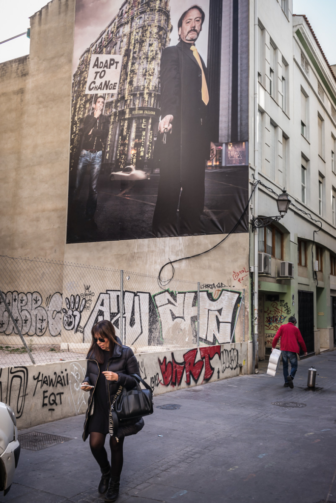 Wandbild mit Stadtgebäude, "ADAPT TO CHANGE"-Schild, Passantin mit Smartphone vorne