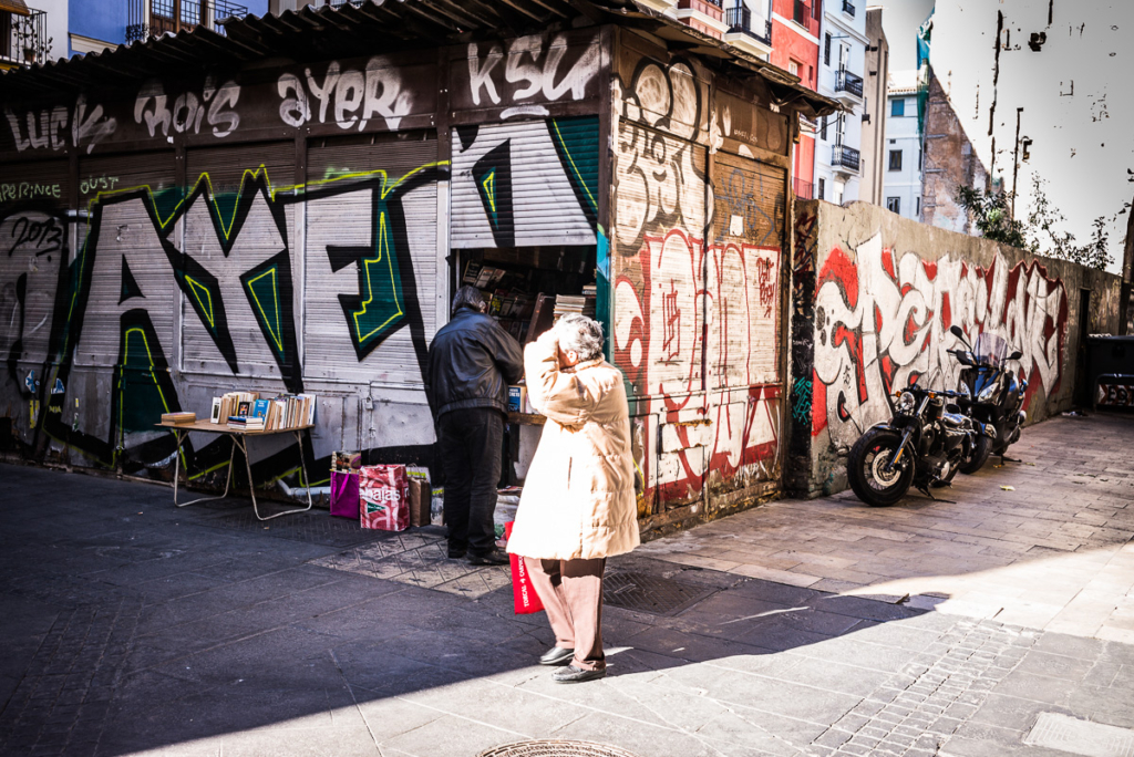 Ältere Frau schützt Augen vor Sonne, steht vor Kiosk mit Graffiti, Bücher ausgestellt, Motorräder.
