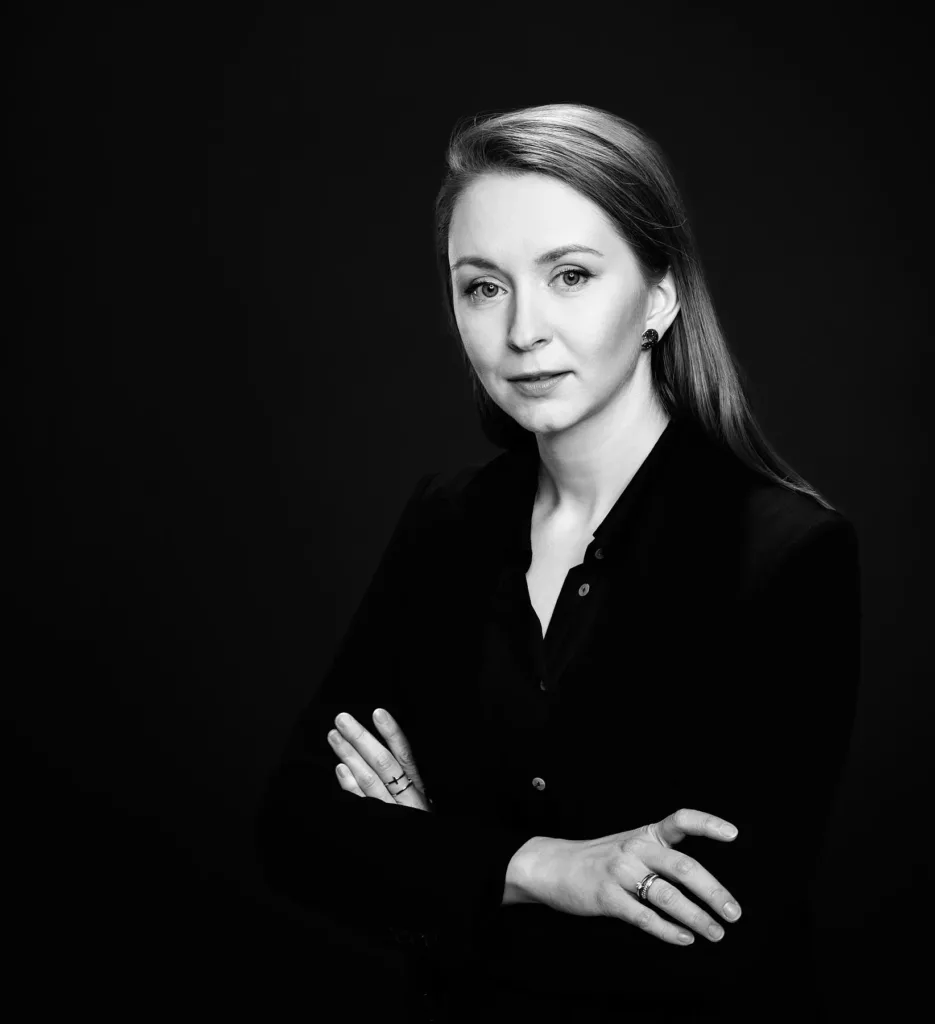 Person im schwarz-weißen Porträt, formell gekleidet, Arme verschränkt, dunkler Hintergrund.