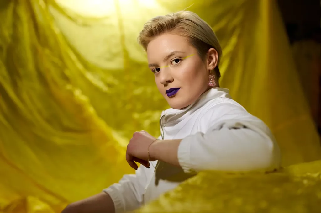 Eine Person mit blondem Haar, Make-up, weißen Oberteil und Ohrringen vor gelbem Hintergrund.