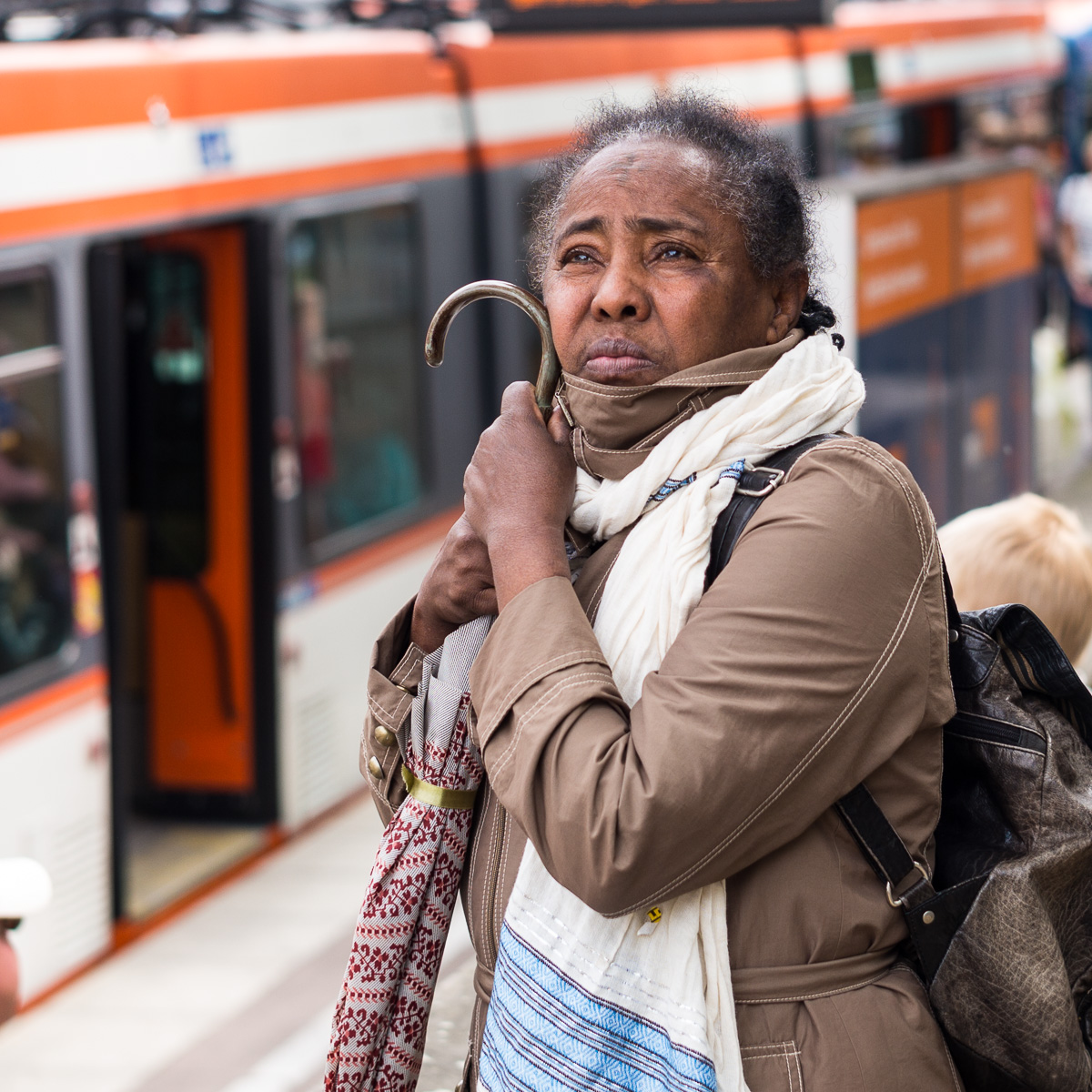 Person am Bahnhof mit beigem Mantel, weißem Schal, Tasche und Regenschirm, Zug im Hintergrund.