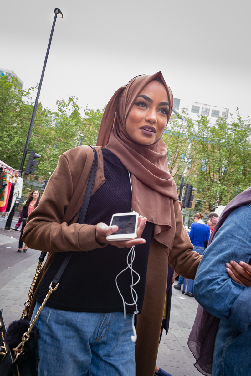Frau in braunem Hijab mit Smartphone und Kopfhörern, Menschen und Bäume im Hintergrund.