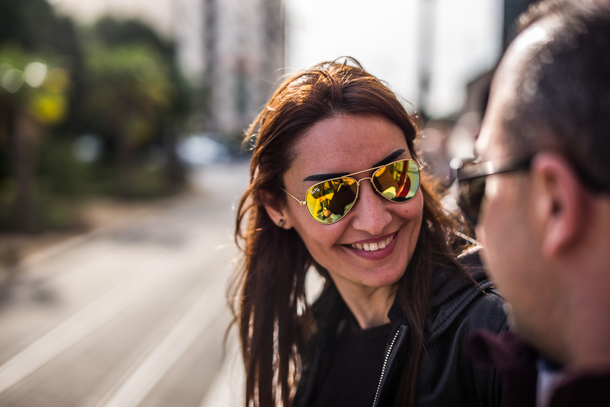 Frau mit Sonnenbrille lächelt Person in städtischer Umgebung an, Straße und Bäume im Hintergrund.